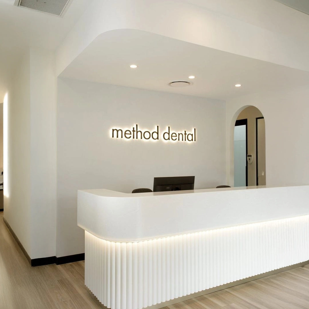 White LED-lit 3D acrylic reception sign displaying “method dental” on a clean white wall in a modern dental clinic interior with curved counters and warm lighting.