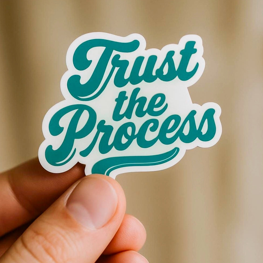 Hand holding a sticker with 'trust the process' text on a light background