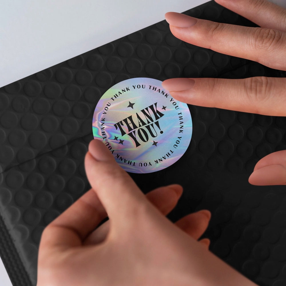 Holographic 'THANK YOU!' sticker held by a hand on a black textured surface