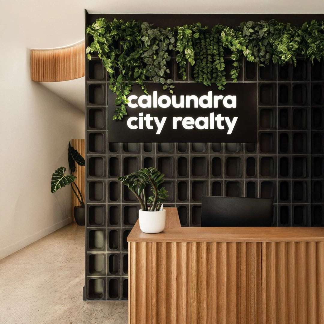 Office reception sign with white 3D letters on a black panel behind a wood reception desk and green wall décor.