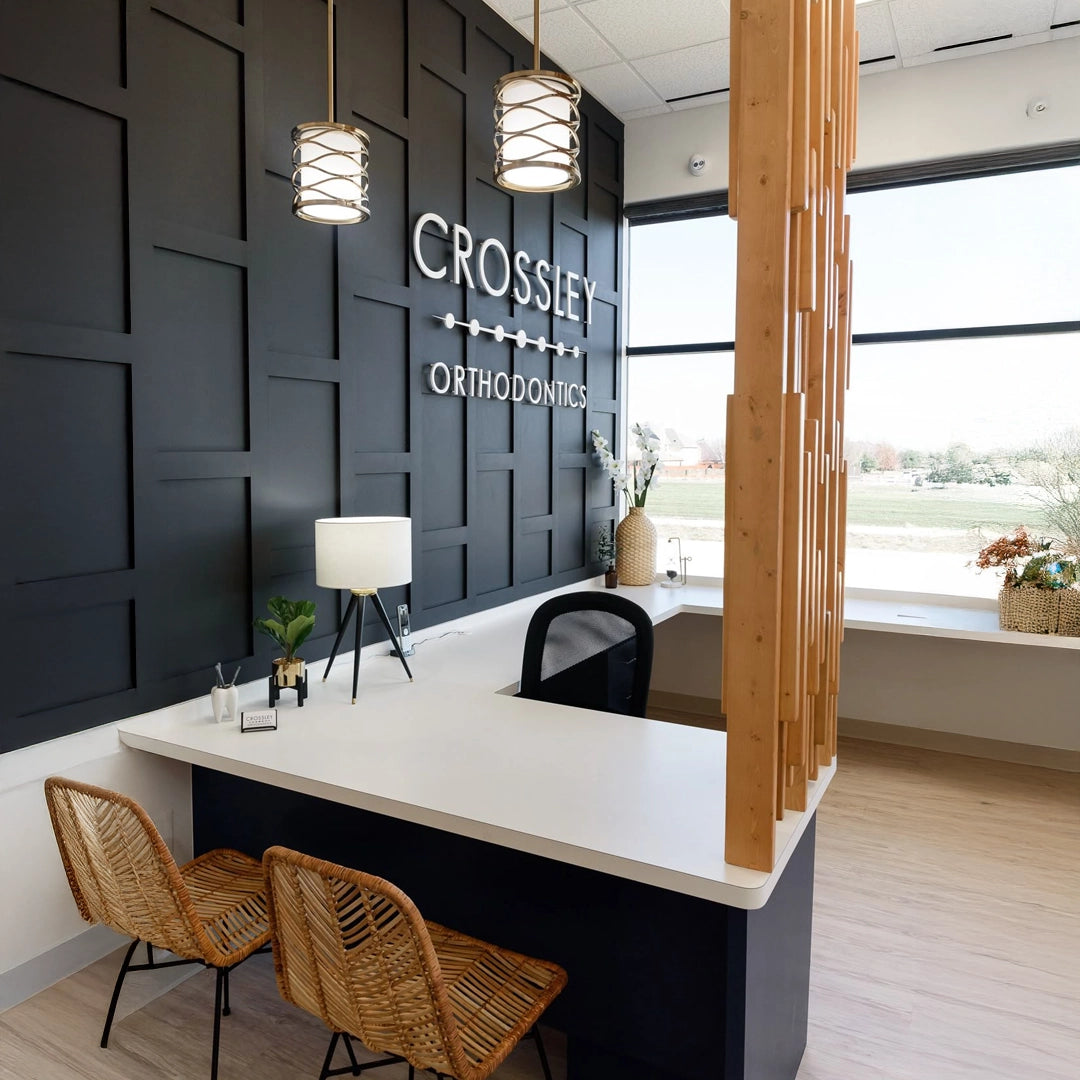 White 3D acrylic wall signage reading “CROSSLEY ORTHODONTICS” mounted on a matte black feature wall in a modern orthodontic clinic reception with rattan chairs and pendant lights.