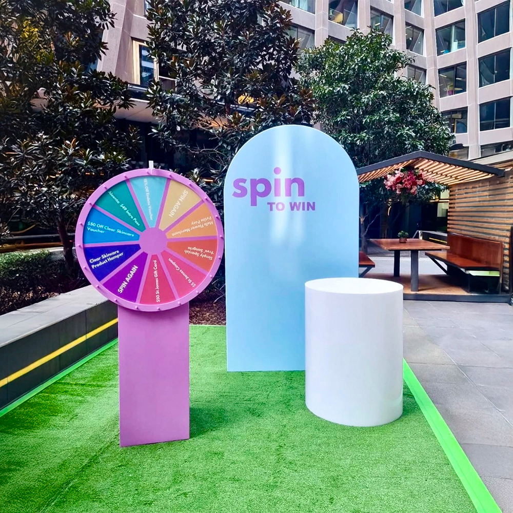 Colorful spinning wheel and 'spin to win' sign in an outdoor setting with trees and buildings in the background.