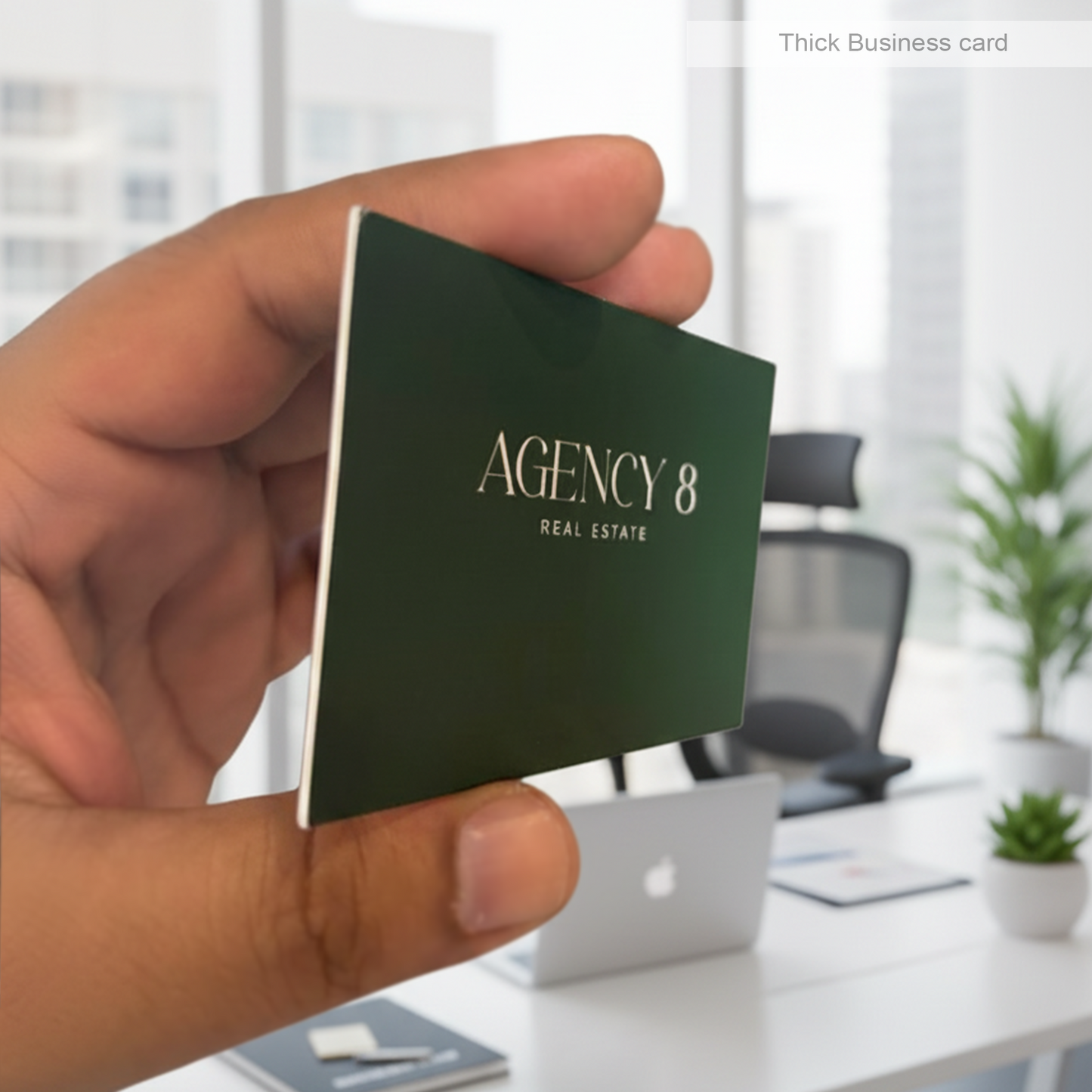 Hand holding a green thick business card with 'Agency 8 Real Estate' printed on it, blurred office background.