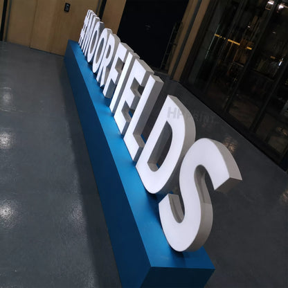 Large 3D led letters spelling 'SHORFIELDS' in white, mounted on a long blue plinth in an indoor industrial or office setting with a gray floor and glass doors in the background.