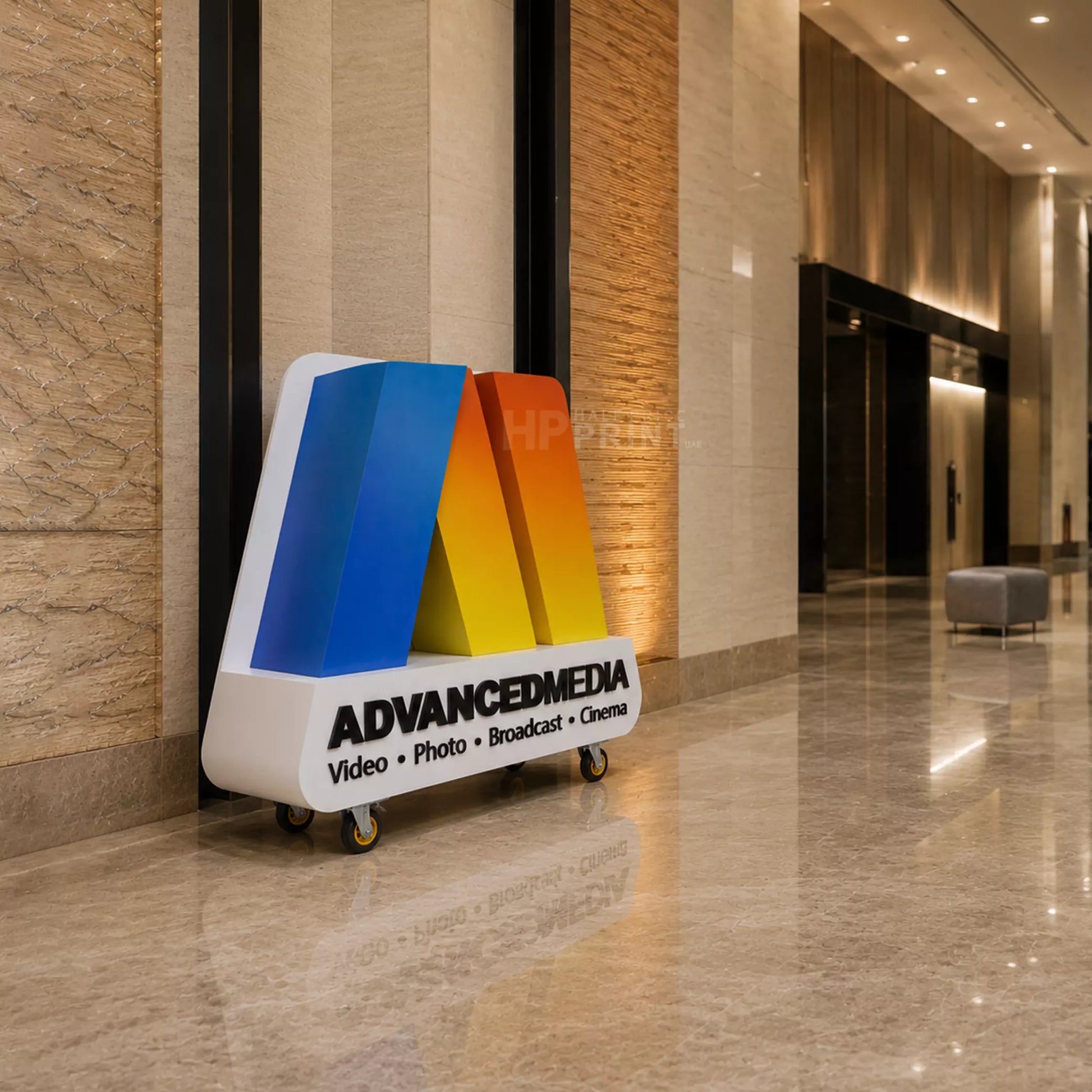 A freestanding modern 3d letters or display stand with a white base, black text reading ADVANCEDMEDIA and smaller text Video Photo Broadcast Cinema, topped with a stylized letterform in blue, yellow, and orange. The stand is photographed in a polished indoor lobby with beige stone walls and floor.