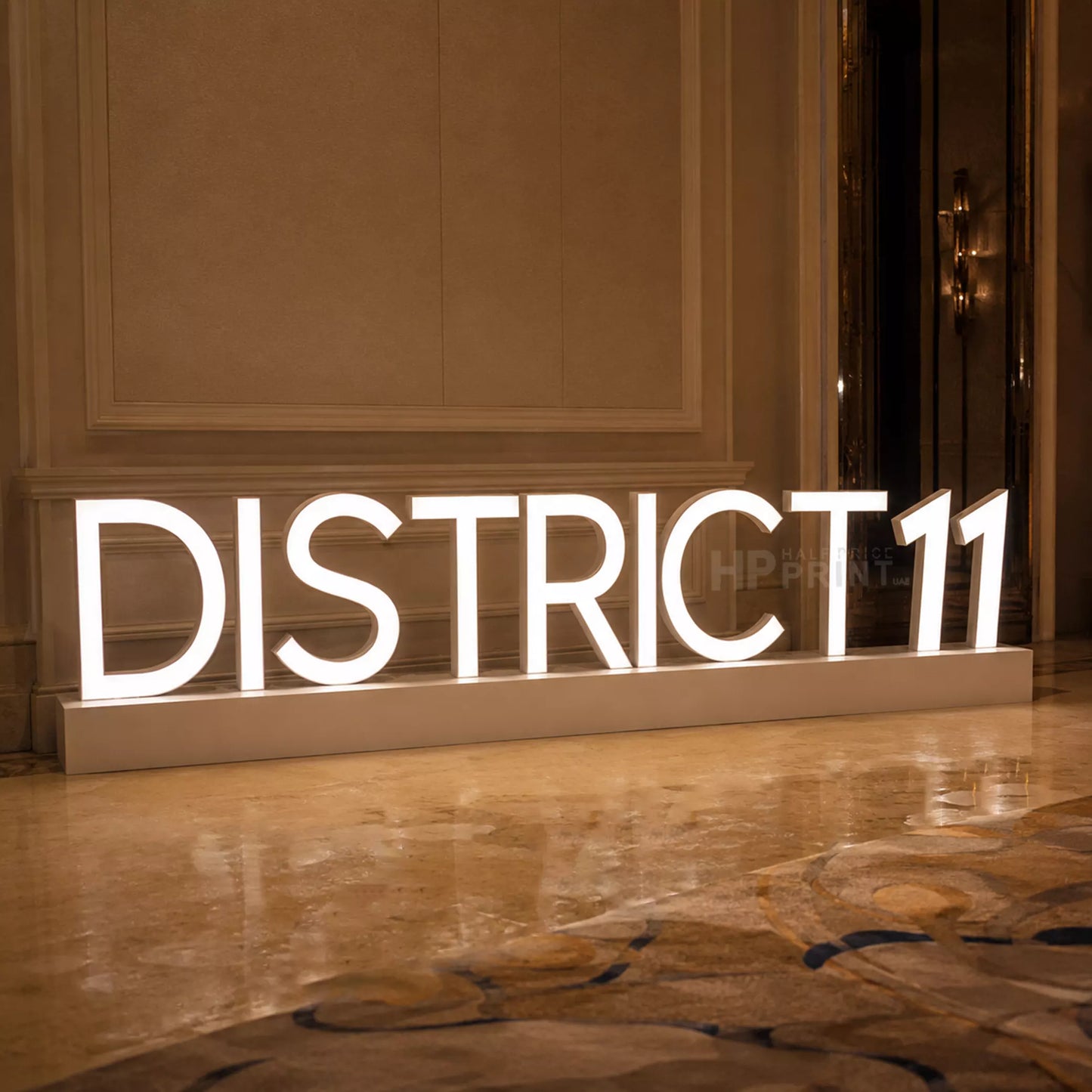 Large illuminated LED freestanding sign displaying 'DISTRICT 11' in white block letters on a rectangular base, placed in an indoor hallway with warm-toned walls and a polished floor.