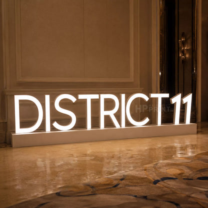 Large illuminated LED freestanding sign displaying 'DISTRICT 11' in white block letters on a rectangular base, placed in an indoor hallway with warm-toned walls and a polished floor.