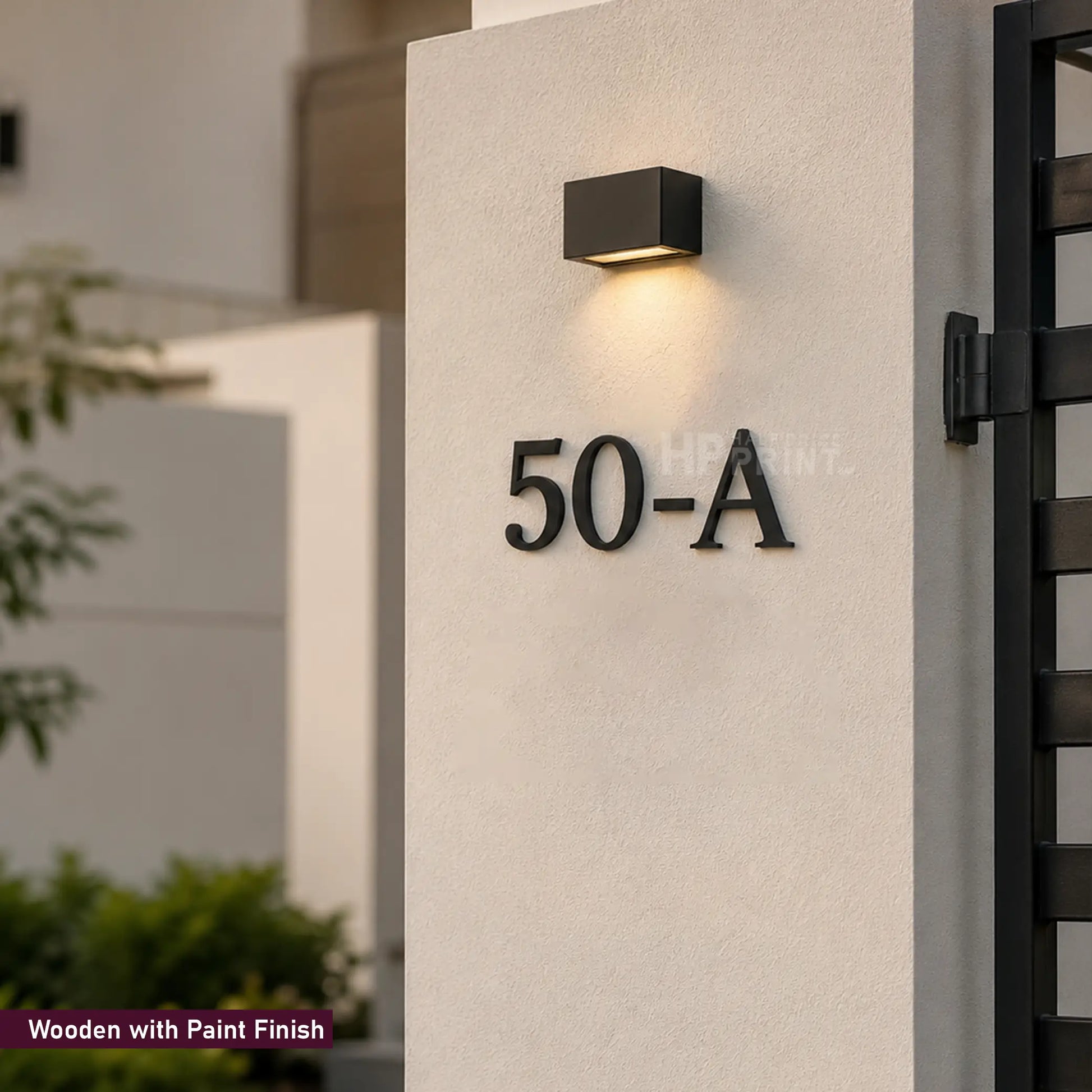 Black metal house number 50-A mounted on a textured off-white stucco pillar, with a modern black square wall sconce above it. The image shows a small text overlay in the bottom-left corner reading 'Wooden with Paint Finish'.