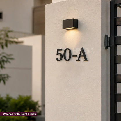 Black metal house number 50-A mounted on a textured off-white stucco pillar, with a modern black square wall sconce above it. The image shows a small text overlay in the bottom-left corner reading 'Wooden with Paint Finish'.