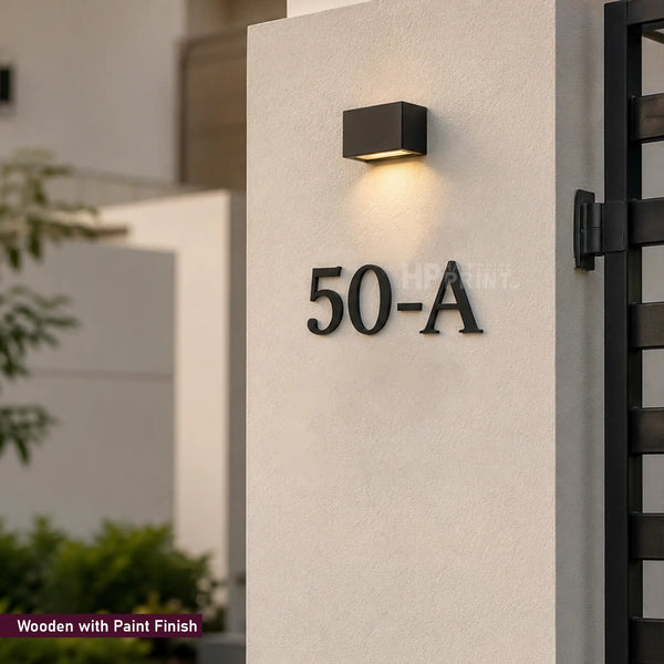 Black metal house number 50-A mounted on a textured off-white stucco pillar, with a modern black square wall sconce above it. The image shows a small text overlay in the bottom-left corner reading 'Wooden with Paint Finish'.