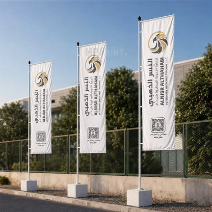 Three vertical white flag banners mounted on poles with concrete bases, displayed outdoors on a sunny day. The flags feature a gold and black circular logo and the text 'AL MISWA ALTHAHABI INTERNATIONAL BEACONS SYSTEMS, LLC' in English and Arabic.