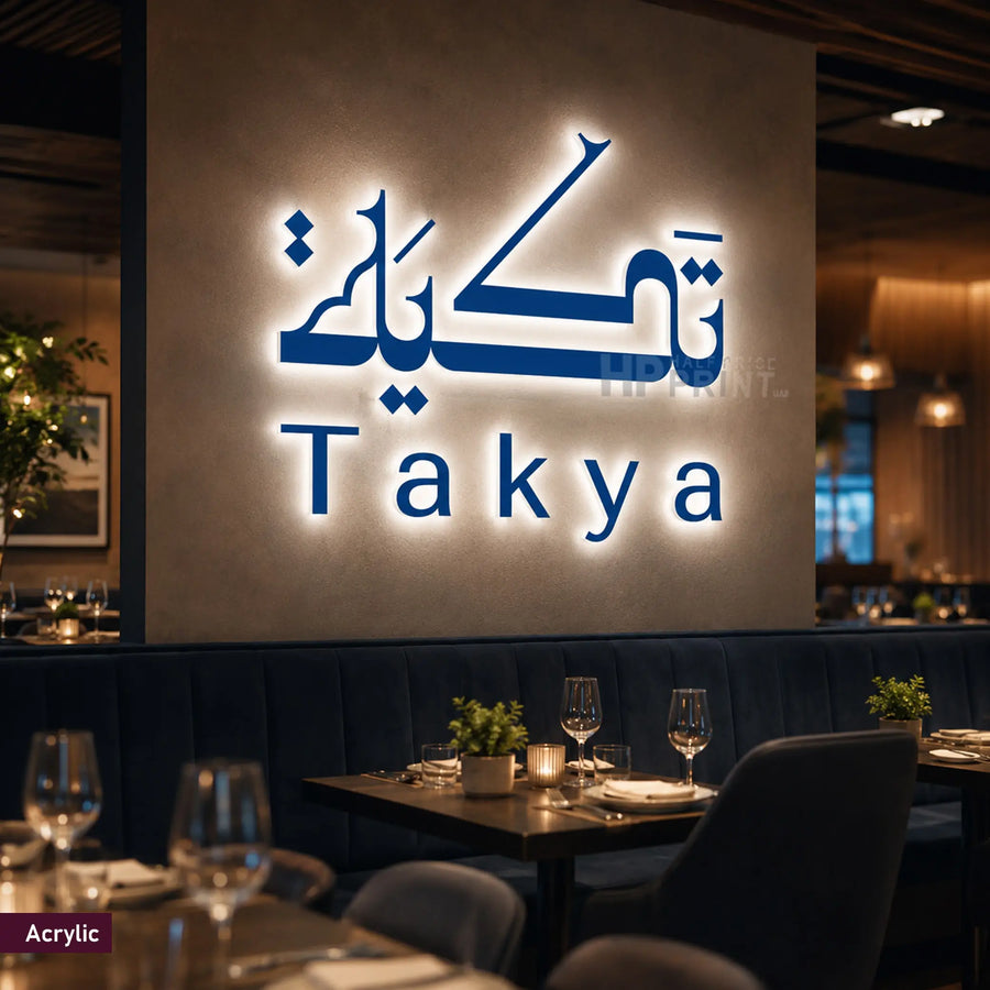 A large illuminated acrylic wall sign displaying the brand name 'Täka' in stylized blue Arabic script with the word 'Täka' underneath, mounted on a warm beige wall in a restaurant setting with tables, chairs, and wine glasses visible in the foreground.