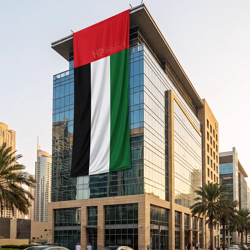 A large vertical building flag of the United Arab Emirates hangs from the side of a modern glass office building in an urban setting. The flag features four equal vertical bands of red, black, white, and green, with the UAE coat of arms printed near the hoist side.