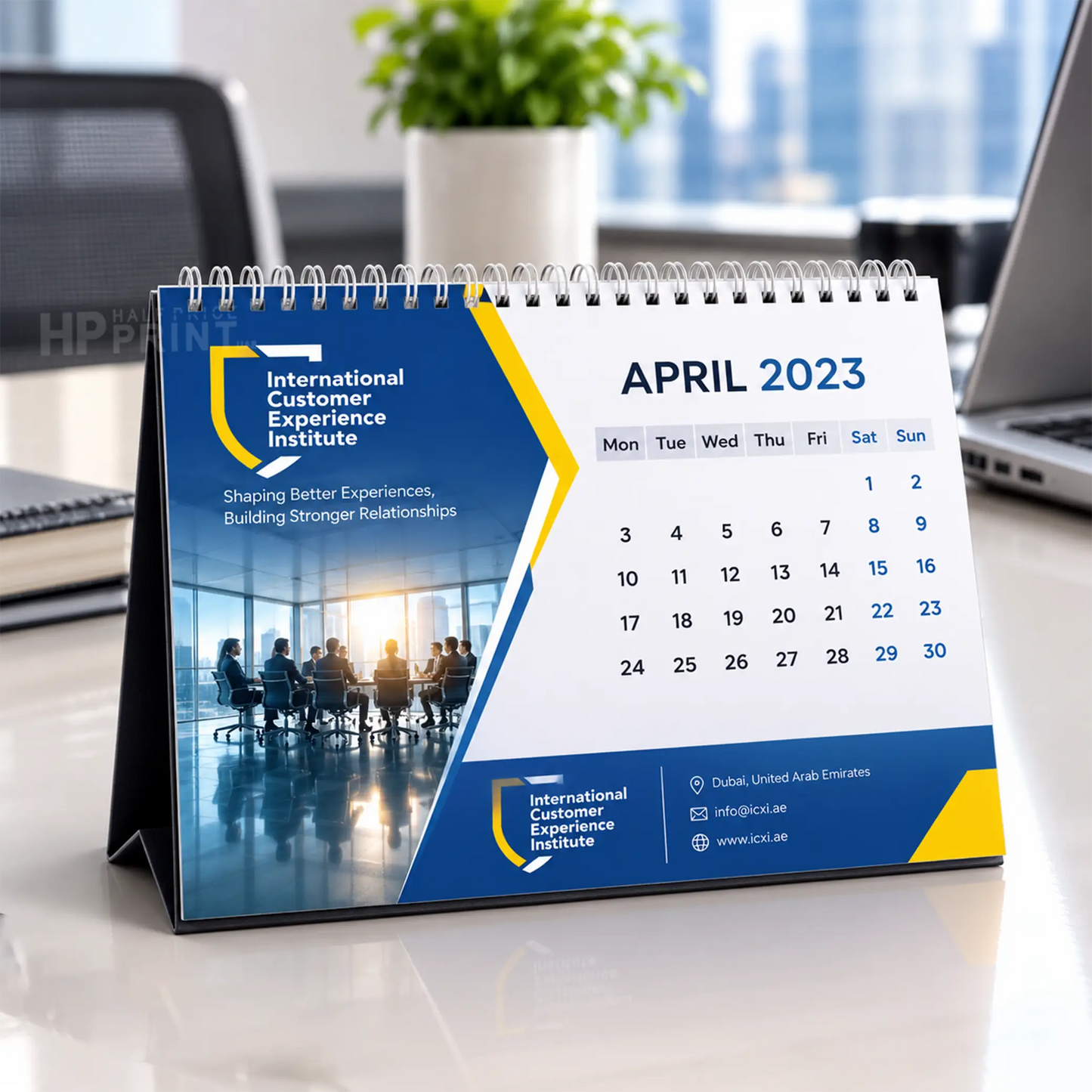 A spiral-bound desk calendar standing on a white surface, printed with the International Customer Experience Institute branding and a April 2023 calendar layout. The calendar has a blue, white, and yellow design with office imagery on the cover.