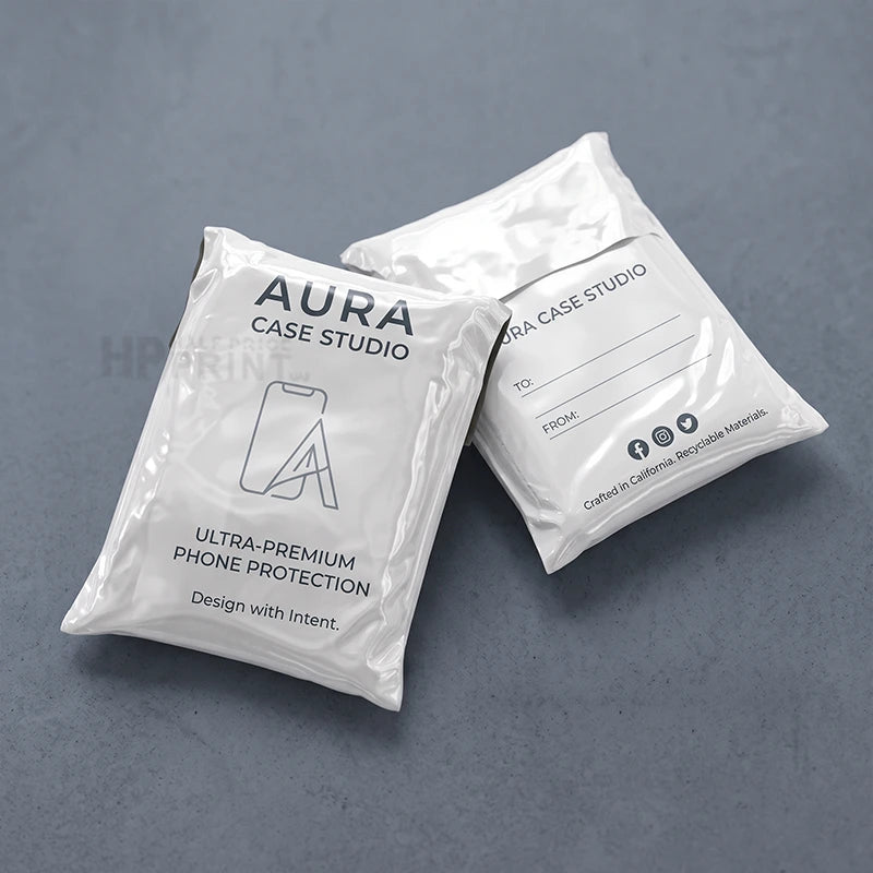 Two white compostable mailers for phone protection with black printed text reading 'AURA CASE STUDIO' and 'ULTRA-PREMIUM PHONE PROTECTION' on a dark gray textured background.