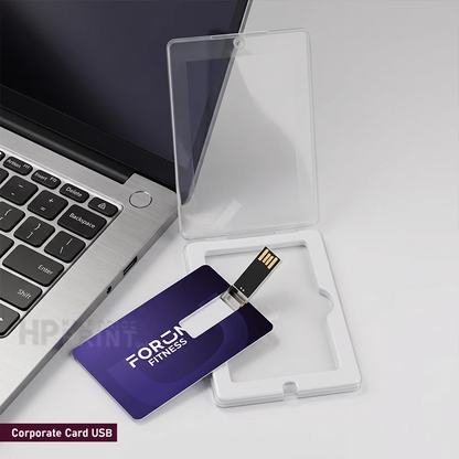 A purple corporate card USB drive with a metal USB connector, shown next to its clear plastic protective case and white storage tray on a light gray surface, with part of a laptop visible in the background.
