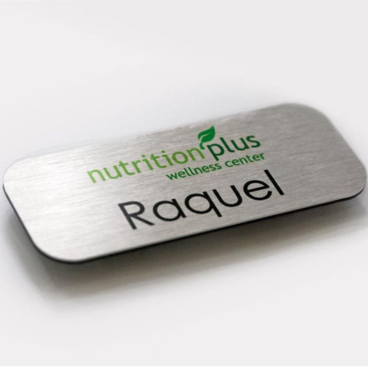 A rectangular name tag with a silver metal finish, green and black text that reads 'nutritionplus wellness center', and a name 'Raquel' printed in black.
