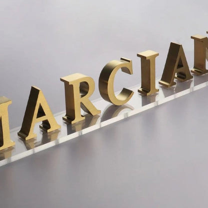Custom metal letters spelling out the name 'MARCIANO' in a gold finish, mounted on a clear acrylic background.