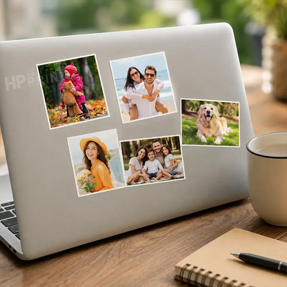 A silver laptop on a wooden desk with five custom photo stickers applied to the back, alongside a mug, notebook, and pen in a blurred outdoor setting.