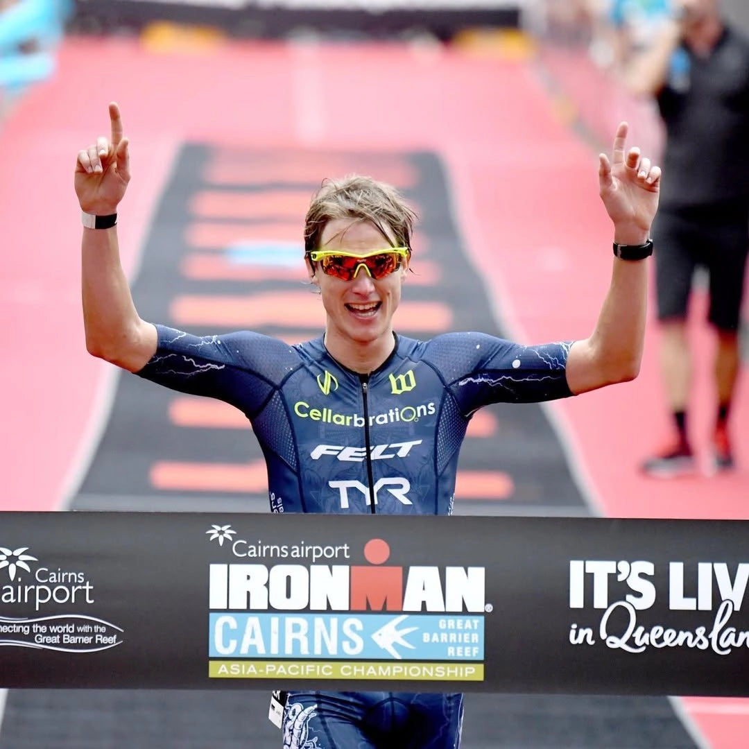 A person celebrating at the finish line of a race wearing sunglasses and a blue triathlon suit with the Ironman logo.