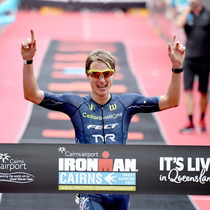 A person celebrating at the finish line of a race wearing sunglasses and a blue triathlon suit with the Ironman logo.