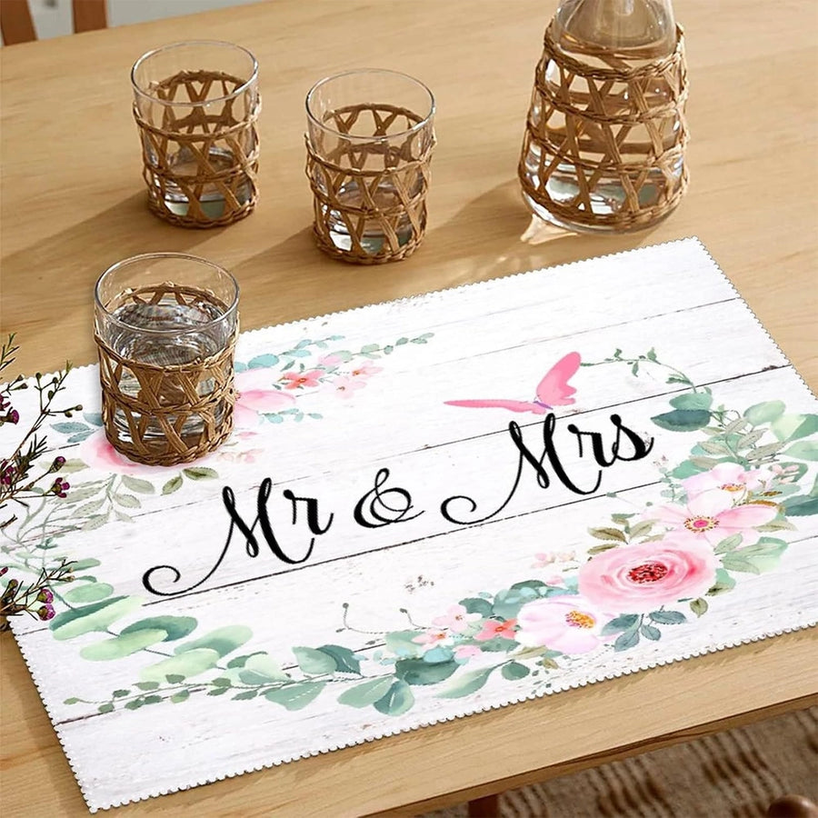 Rustic floral table mat on a light wood table with the text “Mr & Mrs,” surrounded by greenery and pink flowers, with glass tumblers wrapped in wicker holders.