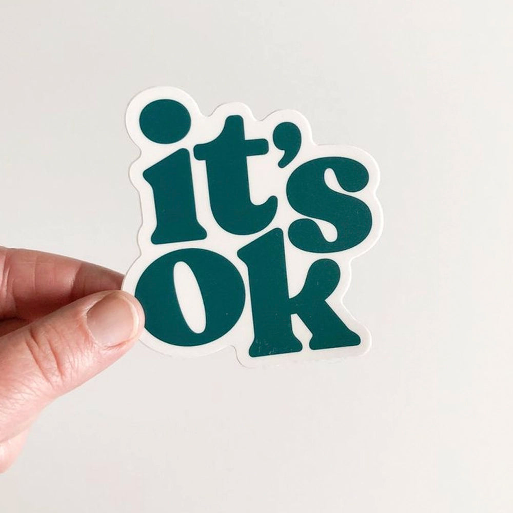 Hand holding a sticker with 'it's ok' text on a light gray background