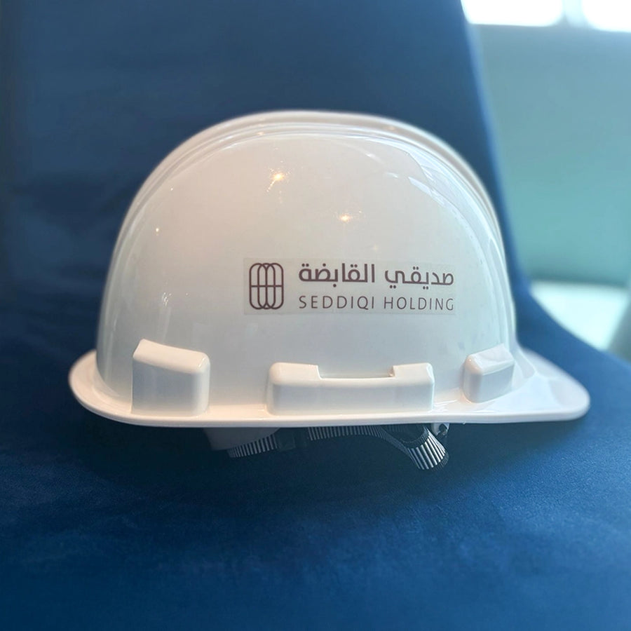 White construction hard hat with the Seddiqi Holding logo and Arabic text printed on the front, resting on a blue chair.