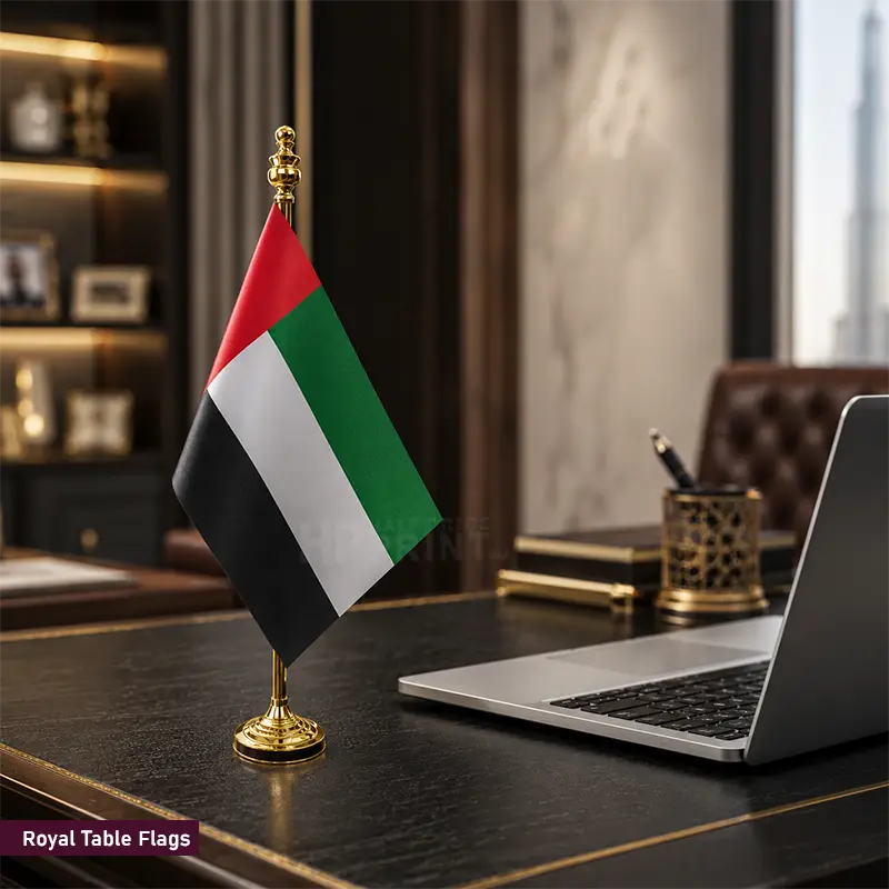 A miniature United Arab Emirates desk flag on a gold-colored stand, displayed on a dark wooden desk next to a laptop in a modern office setting.