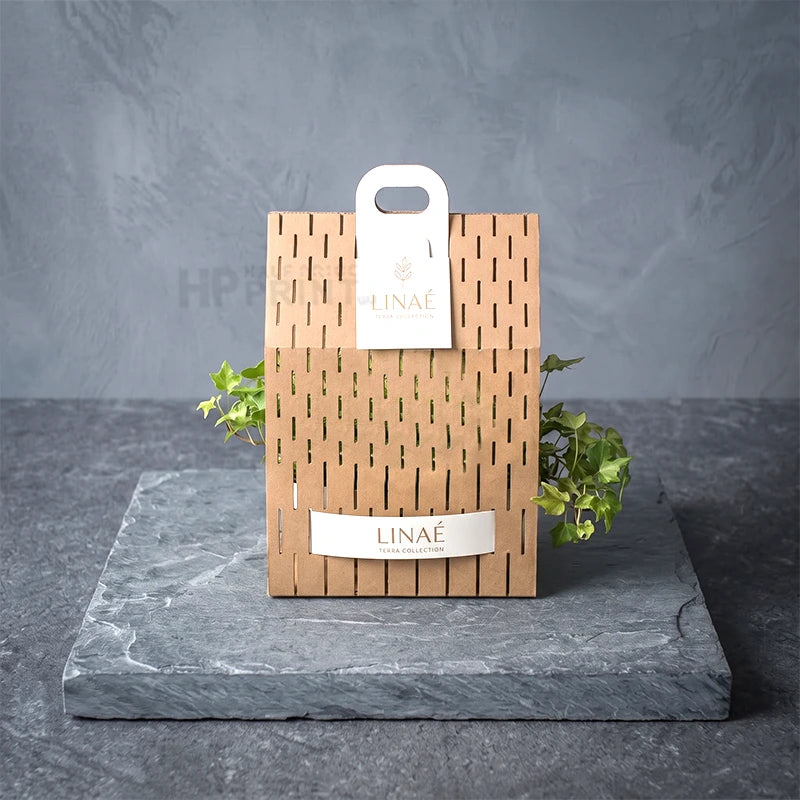 A Linaé branded kraft paper gift die cut box with a slatted front and white hanging tag, displayed on a gray stone slab against a textured gray background with small green plants.