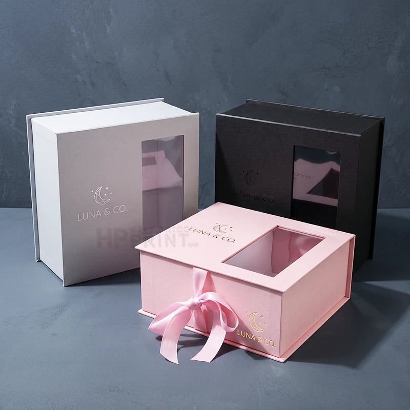 Three custom branded LUNA & CO. gift die-cut boxes with clear viewing windows, shown in white, black, and pink with a pink ribbon, on a textured gray background.