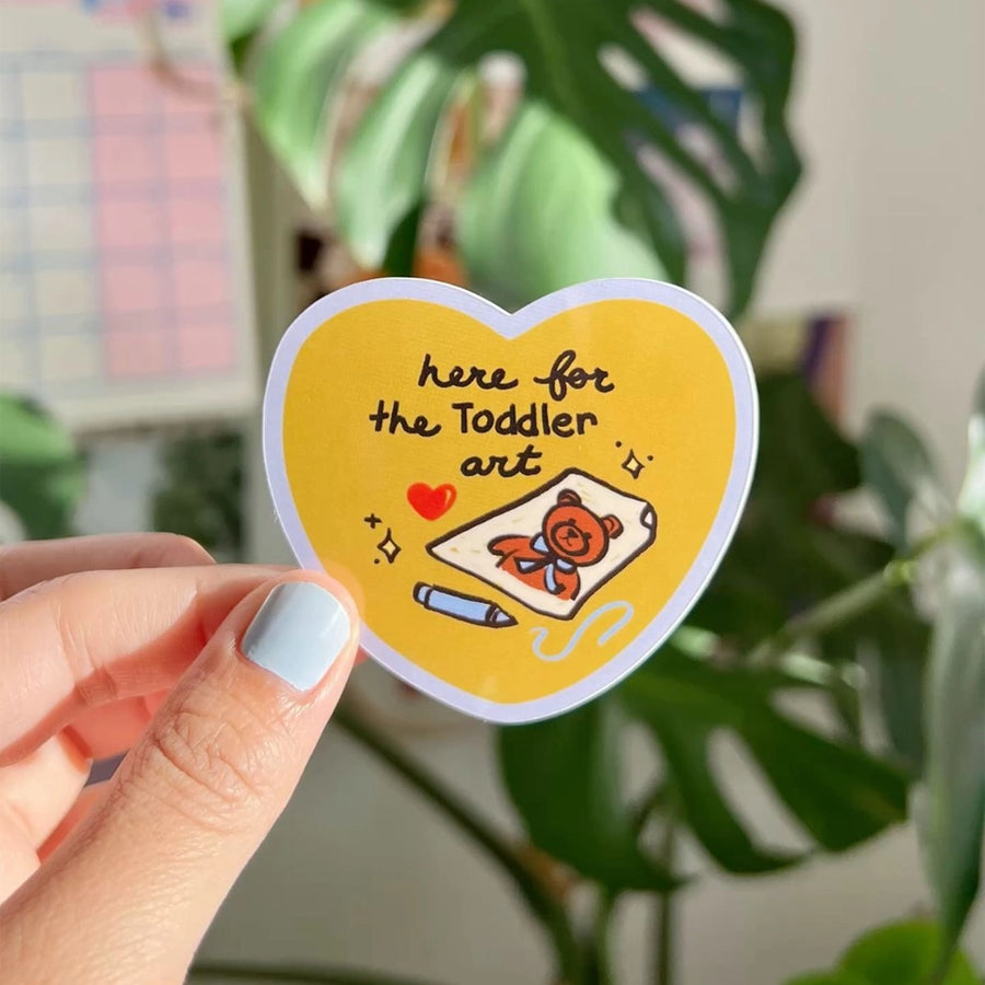 Heart-shaped yellow sticker reading “here for the toddler art,” featuring a crayon drawing of a teddy bear, held in a hand with green houseplants in the background.