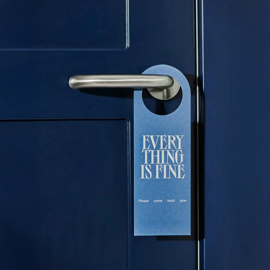A blue door hanger with white text 'EVERYTHING IS FINE' printed on it, hanging on a dark blue door.