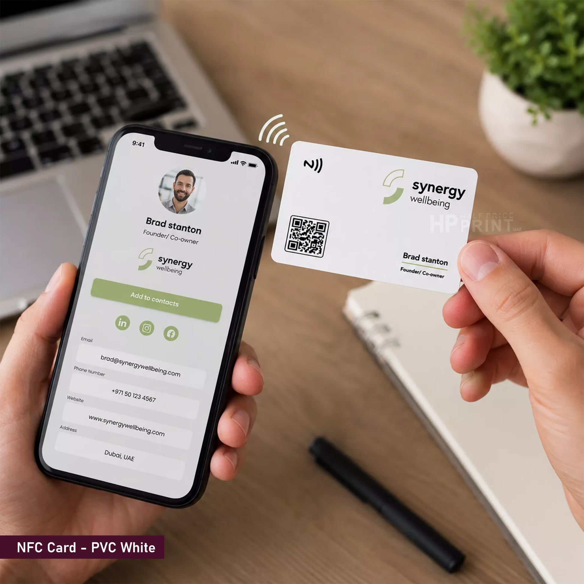A person holds a smartphone and a white NFC-style business card labeled 'synergy wellbeing' with a QR code and name profile. The scene is set on a wooden desk with a laptop, notebook, and plant in the background, and the phone screen matches the card design.