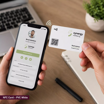 A person holds a smartphone and a white NFC-style business card labeled 'synergy wellbeing' with a QR code and name profile. The scene is set on a wooden desk with a laptop, notebook, and plant in the background, and the phone screen matches the card design.