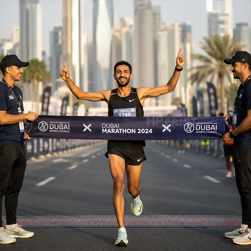 A male marathon runner crosses the finish line at the Dubai Marathon 2024, smiling and raising both hands in celebration. He is wearing a black Nike tank top and shorts, with race officials on either side holding the branded finish line.