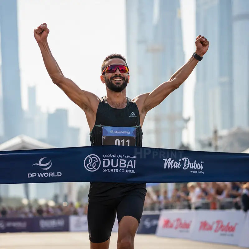 A male athlete celebrating the finish line at a race, with his arms raised in triumph. He is wearing a black athletic singlet and black shorts, with a race bib pinned to his chest.