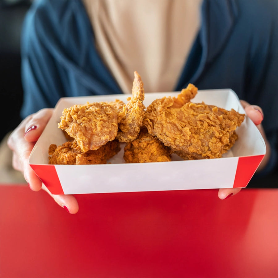 A person holding a red-and-white paper packaging tray filled with crispy fried chicken pieces on a red table.