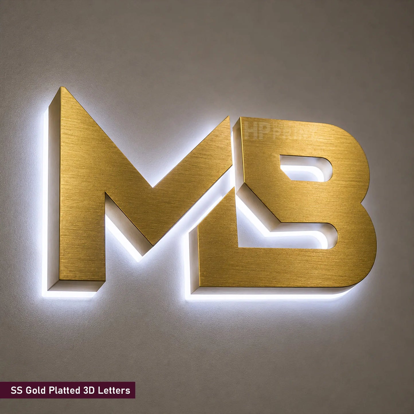 Gold-plated 3D letters 'MB' with integrated LED backlighting, mounted on a neutral gray wall. A small text label in the bottom-left corner reads 'SS Gold Plated 3D Letters'.