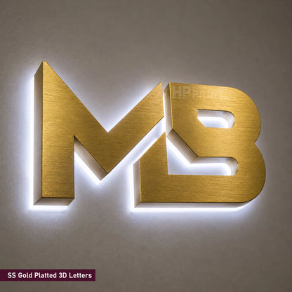 Gold-plated 3D letters 'MB' with integrated LED backlighting, mounted on a neutral gray wall. A small text label in the bottom-left corner reads 'SS Gold Plated 3D Letters'.