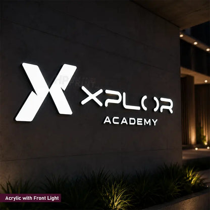 A large illuminated acrylic sign reading 'XPLOR ACADEMY' with a stylized X logo, mounted on a dark building wall at night. The sign is bright white with front-facing lighting, with small plants along the bottom and a faint watermark across the center.