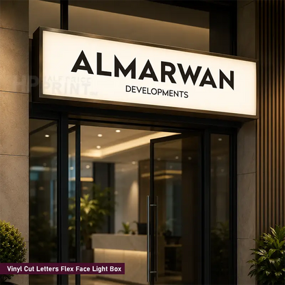A rectangular illuminated light box sign above a glass entrance, displaying the brand name 'ALMARWAN DEVELOPMENTS' in black letters on a warm white face. The sign is mounted on a dark building facade with stone and wood panels, and a small text label at the bottom left reads 'Vinyl Cut Letters Flex Face Light Box'.