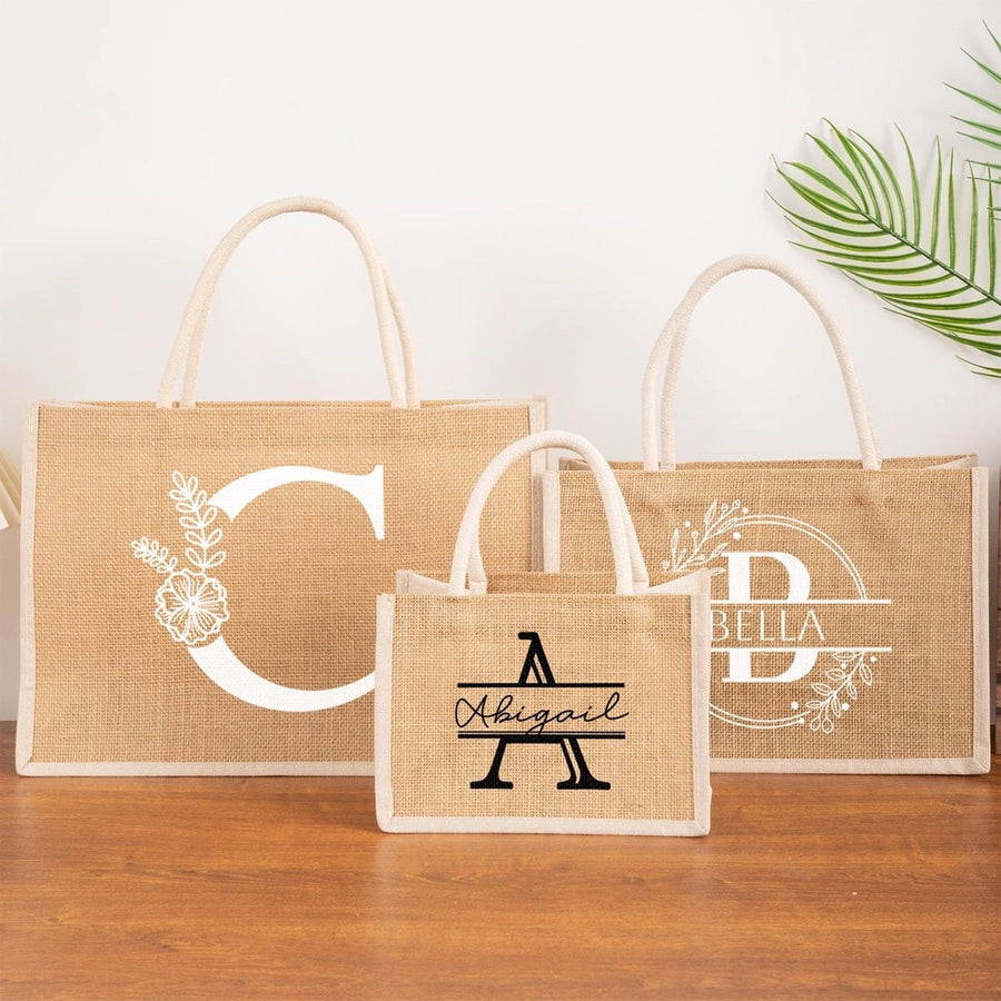 Three natural jute tote bags displayed on a wooden surface, each personalized with a monogram or name in white or black lettering, featuring floral accents and sturdy rope handles.