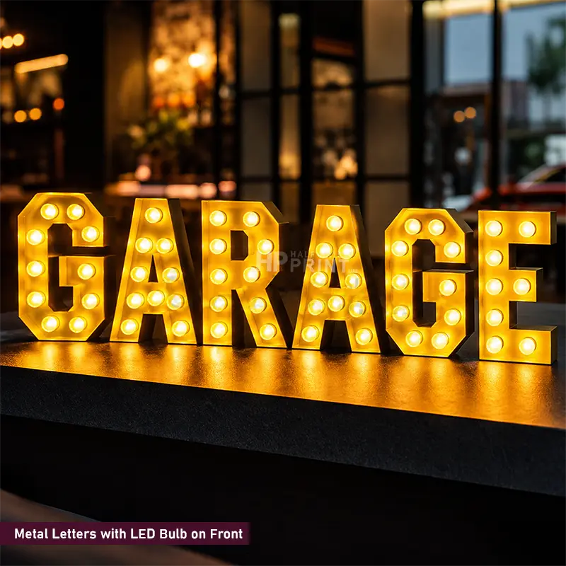 A set of large metal letters spelling "GARAGE" with built-in LED bulbs, glowing warm yellow on a dark surface in an outdoor nighttime setting.