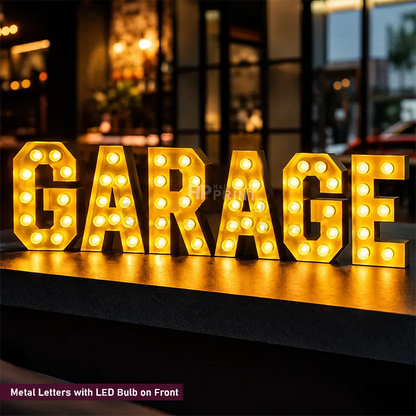 A set of large metal letters spelling "GARAGE" with built-in LED bulbs, glowing warm yellow on a dark surface in an outdoor nighttime setting.