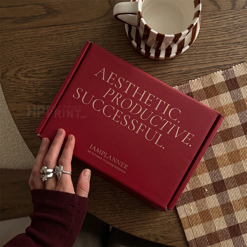 A red mailer shipping box with gold text reading 'AESTHETIC. PRODUCTIVE.