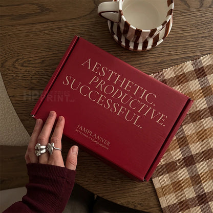 A red mailer shipping box with gold text reading 'AESTHETIC. PRODUCTIVE.