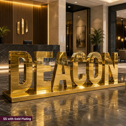 Large gold-plated freestanding letters spelling DEACON placed in a modern lobby or reception area, with polished marble flooring and warm interior lighting in the background.