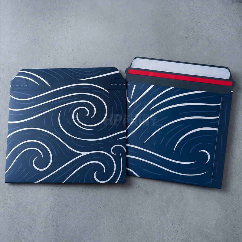 Two navy blue envelope-style mailers with white wave patterns, shown on a gray concrete surface. One envelope is open, revealing a red seal strip and white insert material.