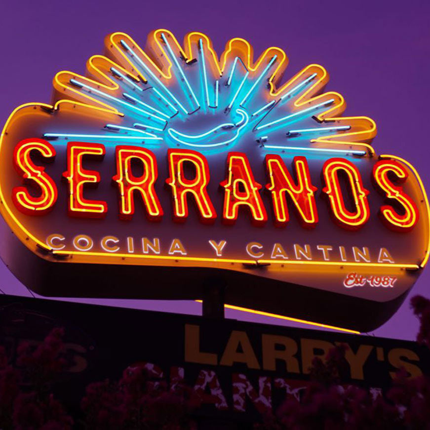 A vibrant neon sign featuring the word 'SERRANOS' in capital letters, illuminated in different shades of blue and red, set against a purple background.